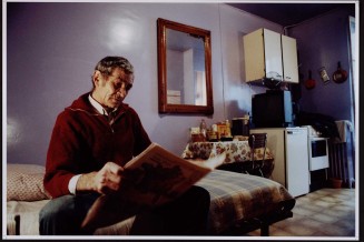 Homme âgé vu de face en biais, lisant son journal, miroir au mur. Guillaume Collanges, "Hôtels meublés - Si tu ne paies pas, tu t’en vas", entre 2000 et 2001, tirages gélatino-argentiques couleur, 41 cm x 62 cm, Collection du Musée national de l'histoire de l'immigration. 2007.59.4
